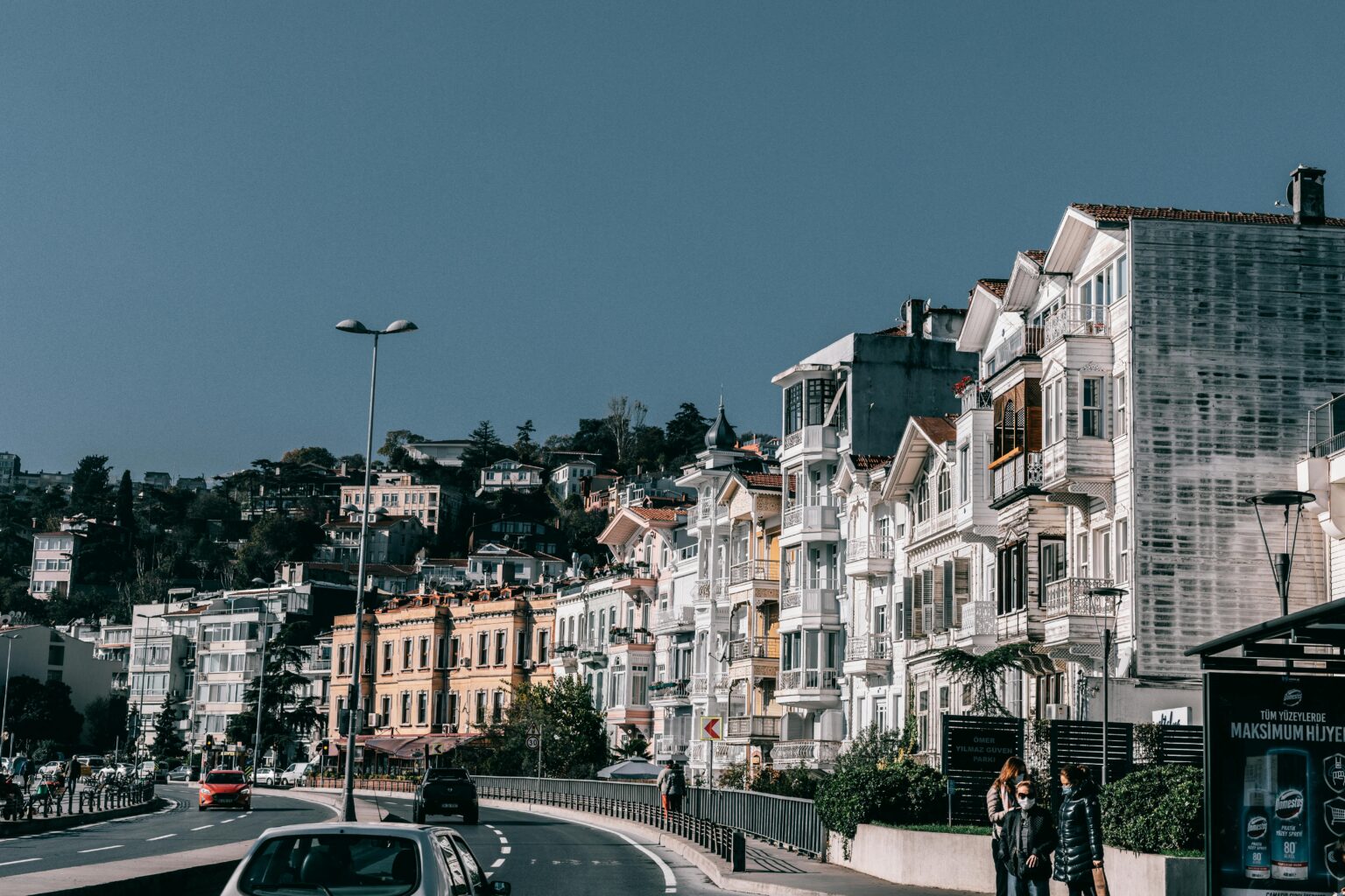Asphalt driveway with cars riding along residential houses located on roadside against blue sky on street with people on bus stop in city